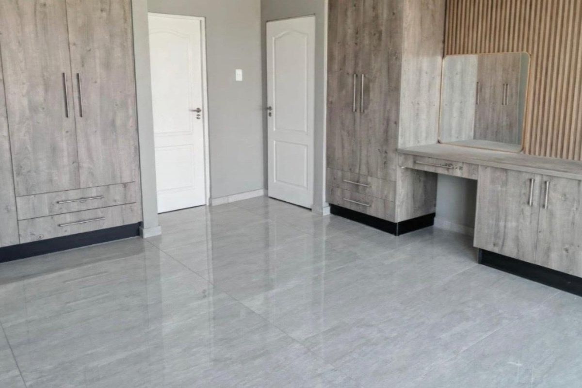 A modern, empty bedroom featuring extensive built-in cupboards, a matching vanity unit, sleek tiled flooring, and decorative slatted wood wall paneling.