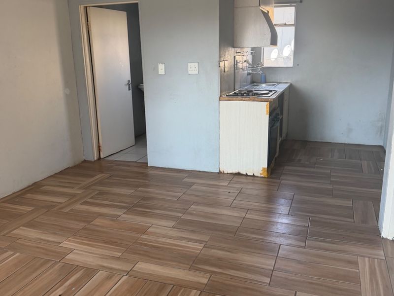 An open-plan living and kitchen area featuring wood-look tiled flooring, a compact kitchenette with built-in appliances, and neutral wall finishes.