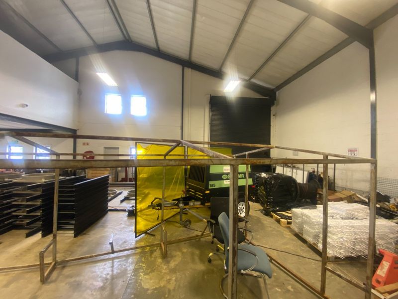 Spacious industrial warehouse unit featuring high ceilings with exposed steel trusses, concrete flooring, and a large roller shutter door for easy access.