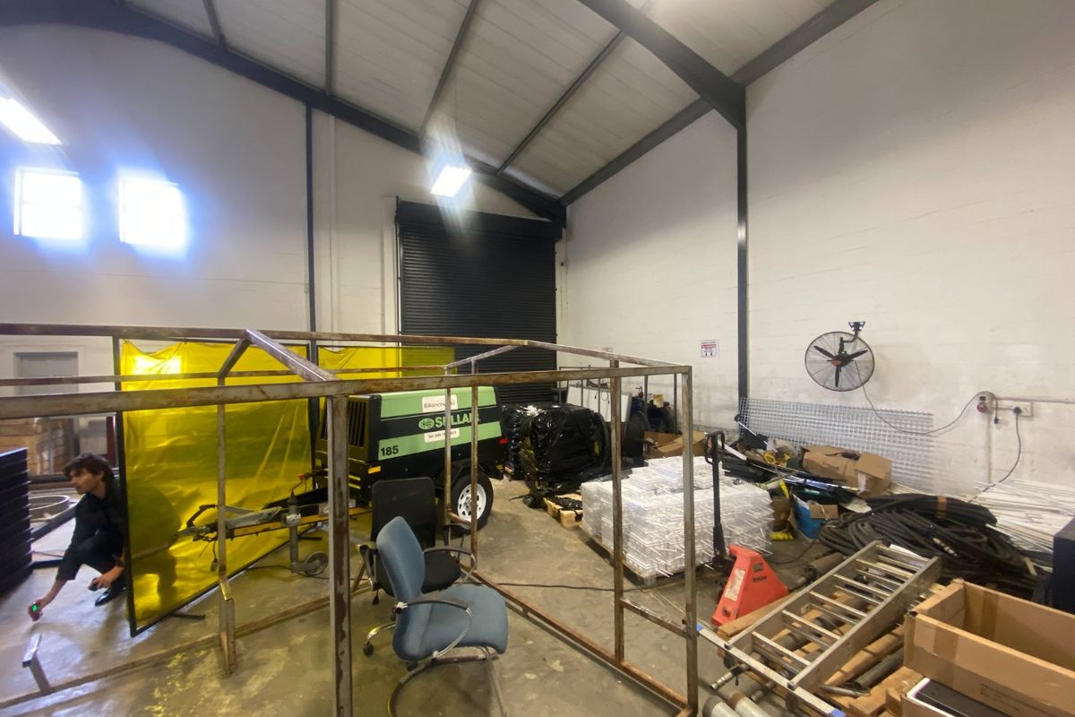 Industrial warehouse utility space featuring high ceilings, exposed steel framing, a large roller shutter door, and concrete flooring suitable for storage or workshop use.