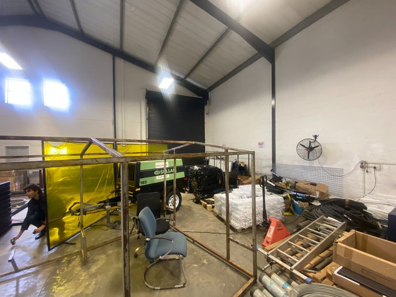 Industrial warehouse utility space featuring high ceilings, exposed steel framing, a large roller shutter door, and concrete flooring suitable for storage or workshop use.
