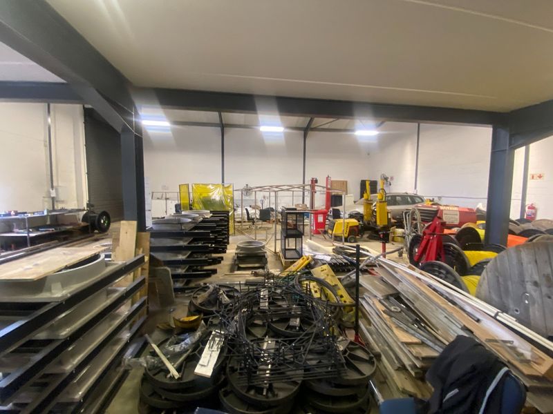 Spacious industrial warehouse area featuring a steel portal frame structure, concrete flooring, and high-bay fluorescent lighting, suitable for manufacturing or storage operations.