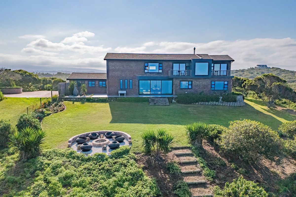 Aerial view of a modern brick farmhouse featuring a spacious lawn, built-in fire pit area, and upper-level balcony overlooking the surrounding landscape.