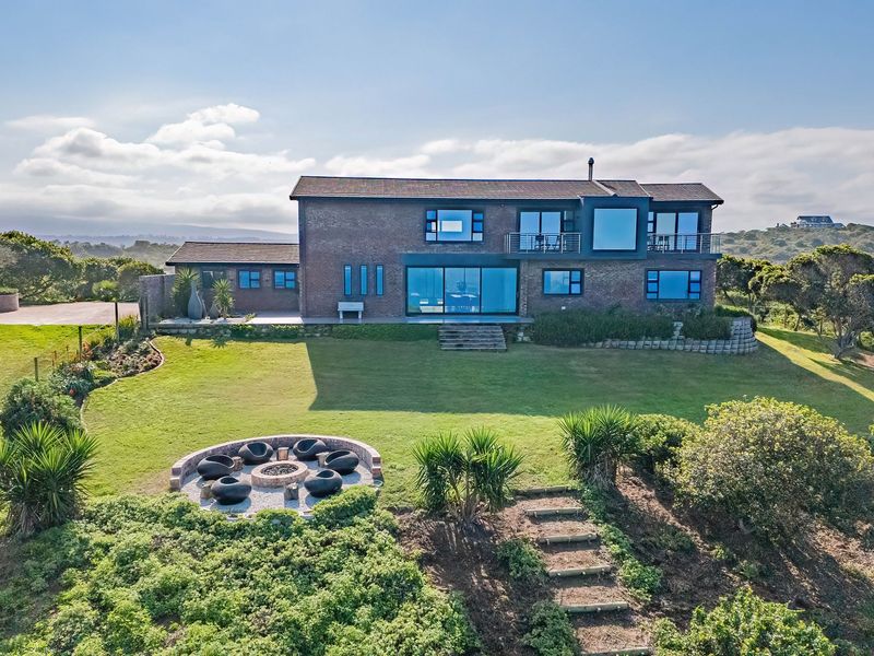 Aerial view of a modern brick farmhouse featuring a spacious lawn, built-in fire pit area, and upper-level balcony overlooking the surrounding landscape.
