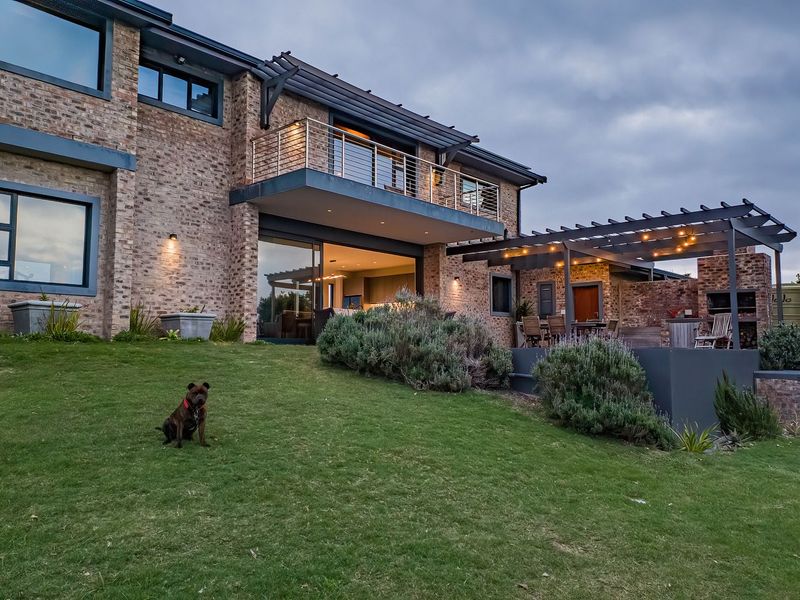 Rear view of a modern brick farmhouse featuring a spacious lawn, a covered patio with a pergola, built-in braai, and an upper-level balcony.