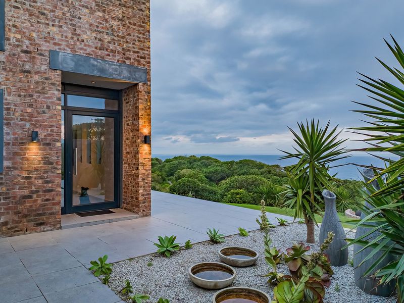 A modern rear exterior featuring exposed brickwork, a large glass entrance door, and a spacious tiled patio overlooking a scenic coastal landscape.