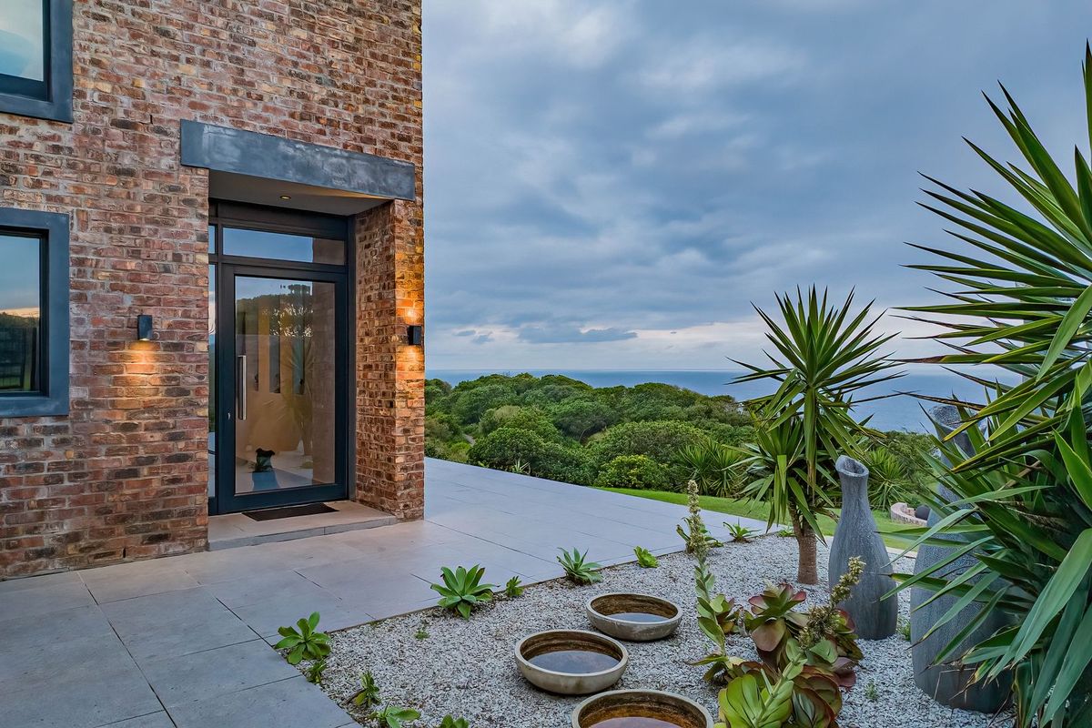 A modern rear exterior featuring exposed brickwork, a large glass entrance door, and a spacious tiled patio overlooking a scenic coastal landscape.