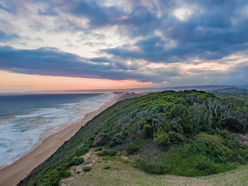 A scenic aerial view of the coastline featuring a sandy beach, ocean waves, and lush coastal vegetation under a dramatic sunset sky.