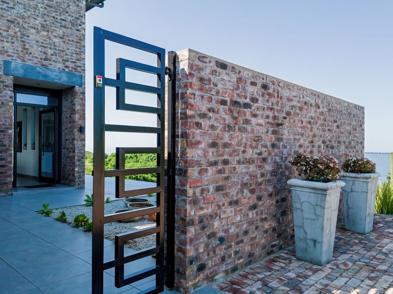 A modern entrance area featuring a striking geometric metal gate, exposed brick walls, and a clean tiled patio leading to the main glass entrance door.