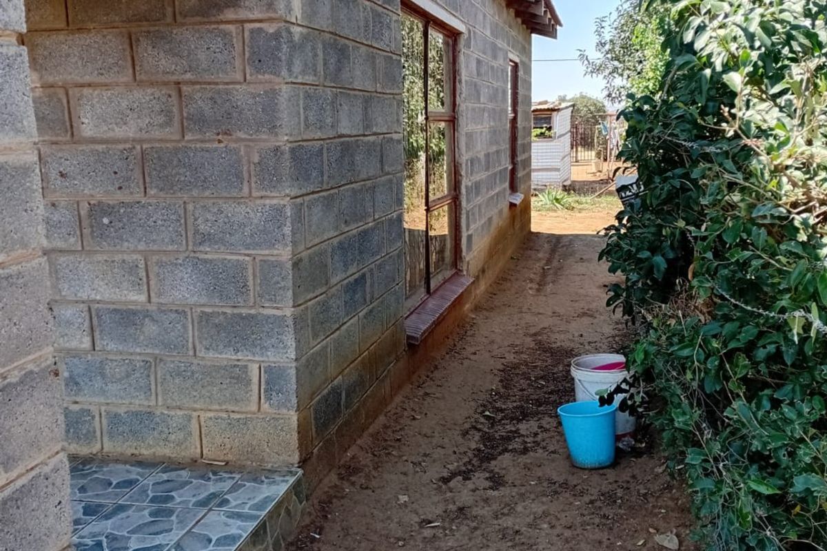 Side view of a house featuring exposed concrete block walls, a small tiled porch area, and metal window frames, set against a natural garden border.