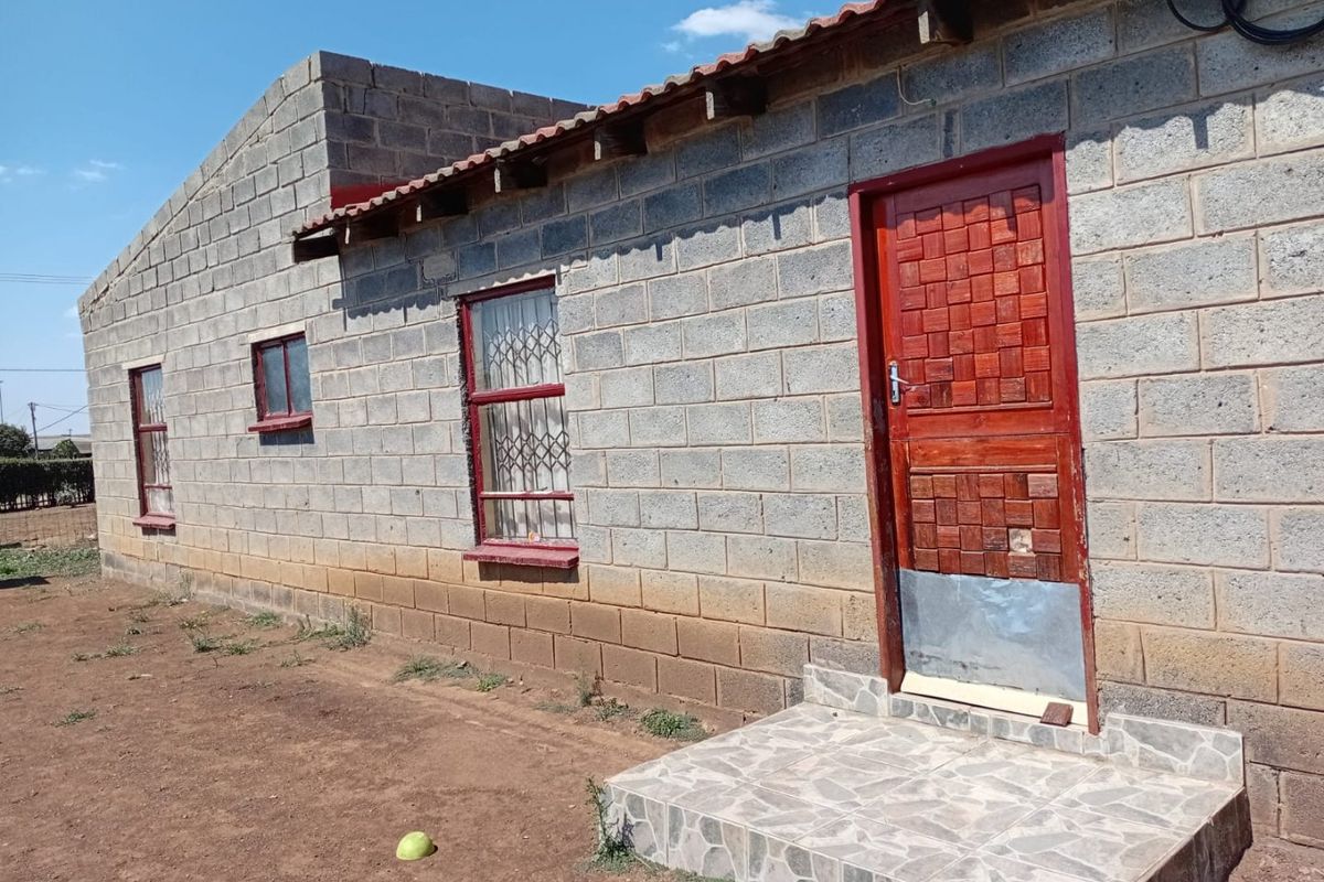 Exterior view of a concrete block house featuring a tiled entrance step, security-fitted windows, and a red wooden door requiring maintenance.