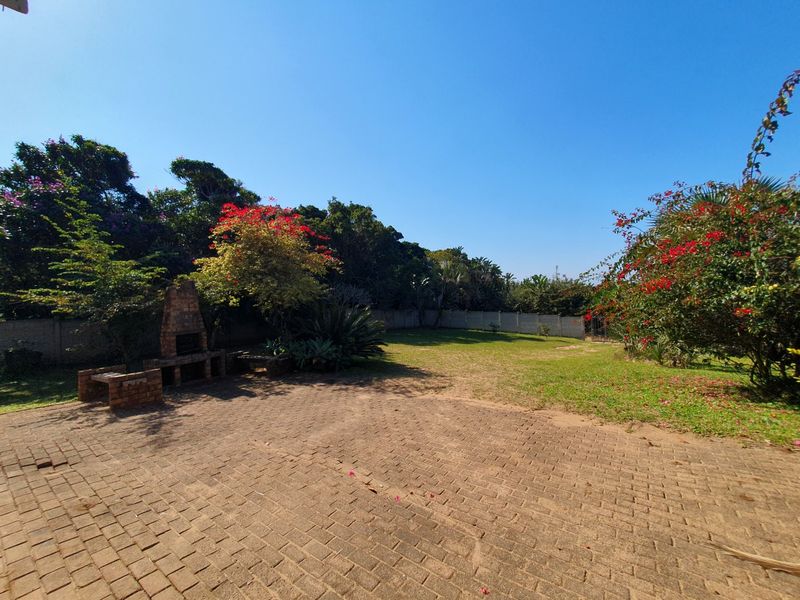 Spacious rear garden featuring a brick-paved patio area with a built-in brick braai, surrounded by established trees and a perimeter wall.