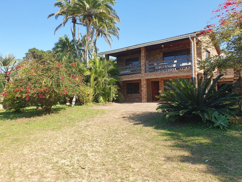 A double-story face brick house featuring an upper-level balcony, paved driveway, and established garden with mature palm trees.