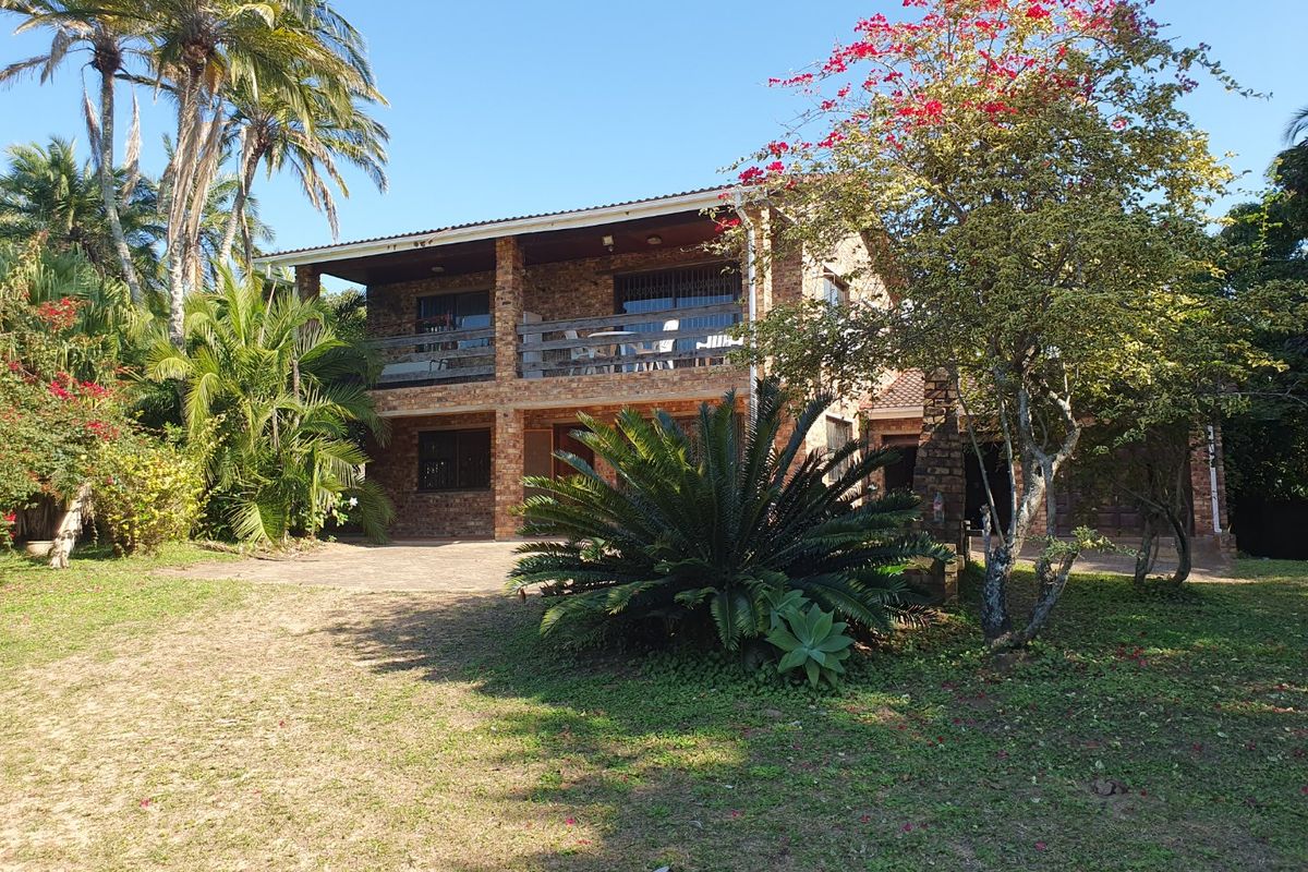 A two-story face brick house featuring a covered upper balcony with timber balustrades, set within a mature landscaped garden with a paved driveway.