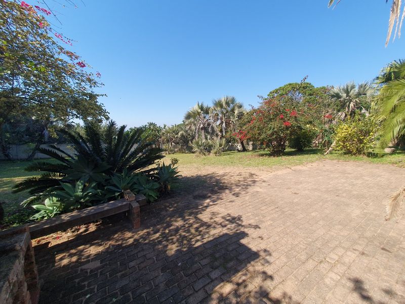 A spacious outdoor garden area featuring a brick-paved patio, a built-in brick bench, and established landscaping with mature trees and shrubs.