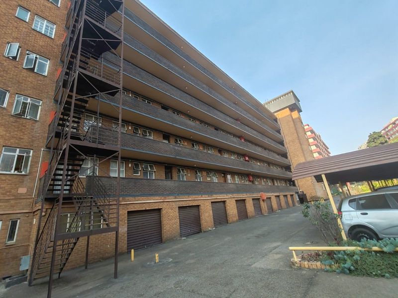 Exterior view of a multi-story brick apartment building featuring an external steel staircase, ground-level garages, and a paved driveway area.
