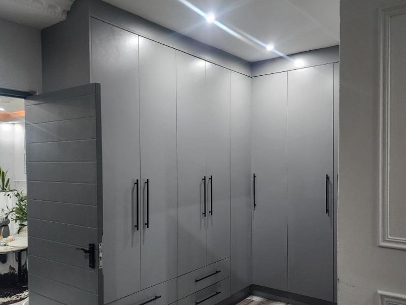 A modern bedroom featuring extensive grey built-in cupboards, recessed downlighting, and high-quality laminate flooring with decorative ceiling cornicing.