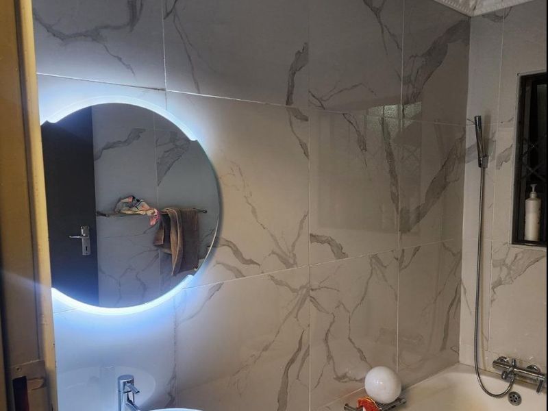 Modern bathroom featuring marble-look wall tiles, a built-in bathtub with handheld shower, a basin, and an illuminated circular mirror.