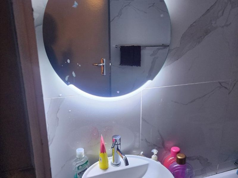A modern bathroom vanity area featuring a vessel sink, chrome faucet, and a contemporary backlit circular mirror against marble-look wall tiles.
