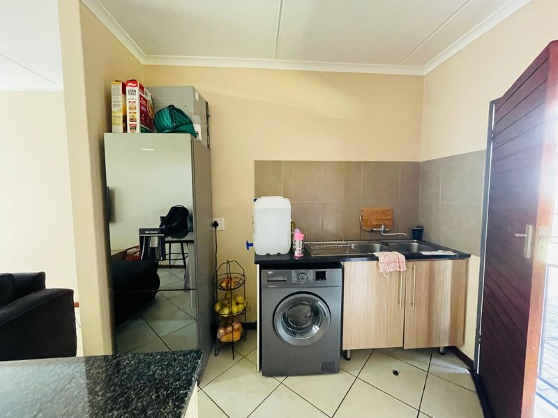 A compact kitchen area featuring tiled flooring, a double bowl sink with tiled splashback, and built-in cabinetry with space for a washing machine.