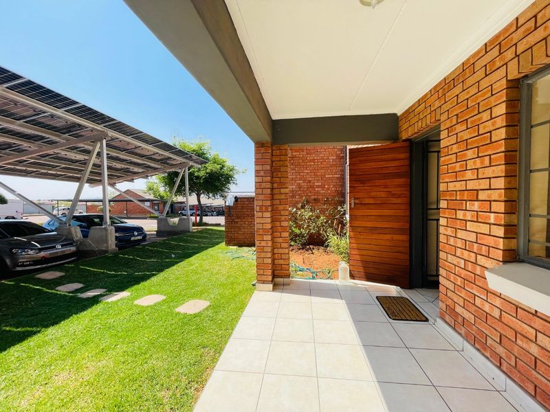 Front entrance area featuring a tiled porch, exposed brickwork, and a timber door, adjacent to a lawn area with a solar-paneled carport.