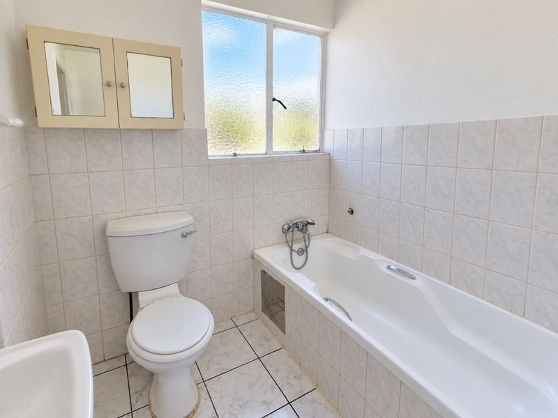 A functional bathroom featuring a standard bathtub, toilet, and wall-mounted medicine cabinet with neutral tiled finishes and natural light from an obscured glass window.
