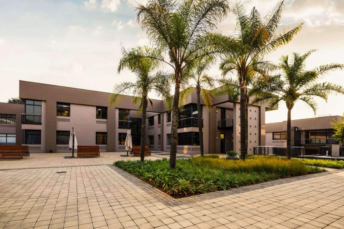 A well-maintained commercial courtyard featuring paved walkways, landscaped garden beds with palm trees, and modern multi-story building facades with balconies.