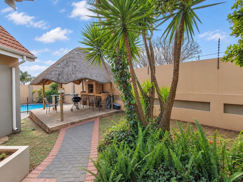 Outdoor entertainment area featuring a thatched lapa with a built-in braai, adjacent to a swimming pool, set within a private garden with paved pathways.