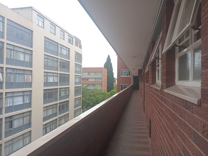 An exterior balcony walkway featuring brick walls, tiled flooring, and a metal balustrade, overlooking surrounding apartment buildings.