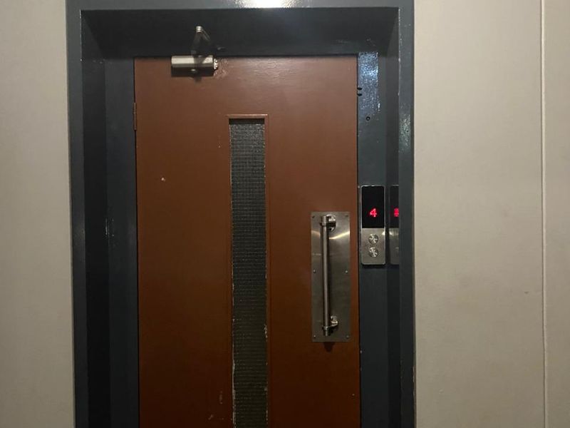 A functional apartment building elevator entrance featuring a brown door with a vertical vision panel and stainless steel hardware.