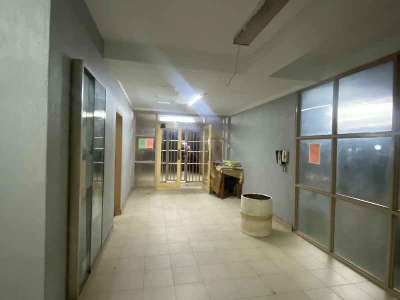 A spacious, tiled hallway featuring a metal security gate, glass partitions, and basic lighting, presenting a functional but dated communal area.