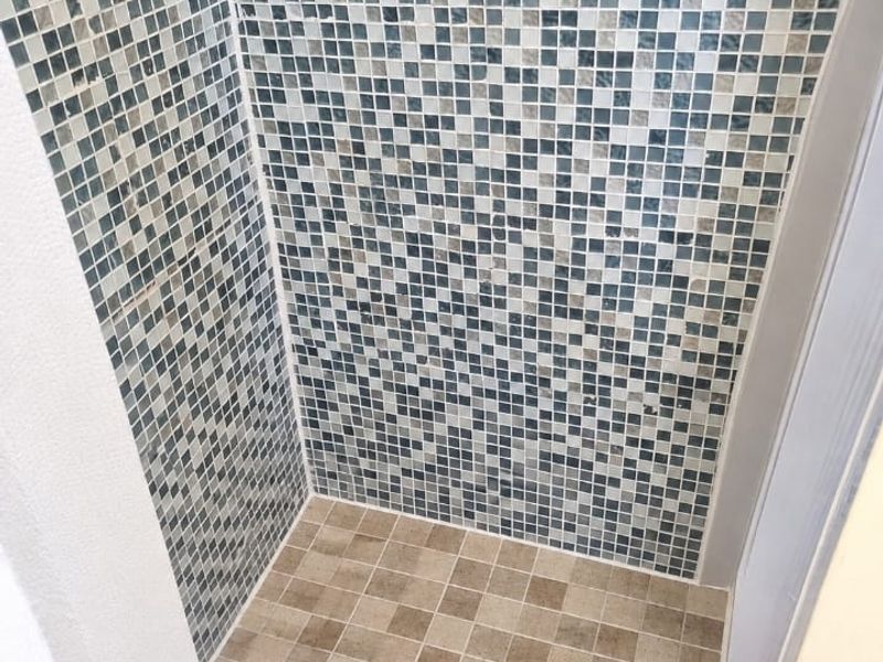 A walk-in shower enclosure featuring decorative mosaic wall tiling and neutral floor tiles with a central drain.