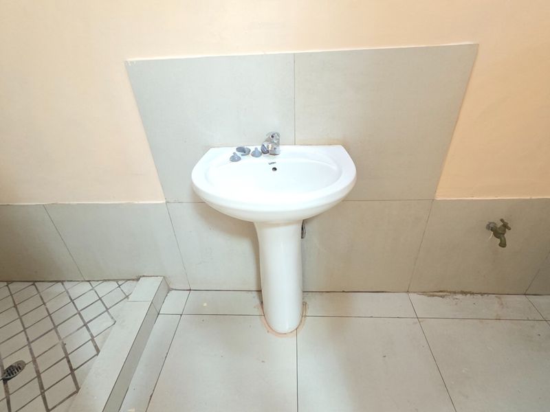 A functional bathroom featuring a white pedestal basin, tiled splashback, and neutral floor tiling, presenting a basic and dated aesthetic.