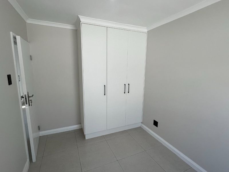 A clean, modern bedroom featuring neutral wall tones, large format floor tiles, and a white built-in wardrobe with contemporary hardware.