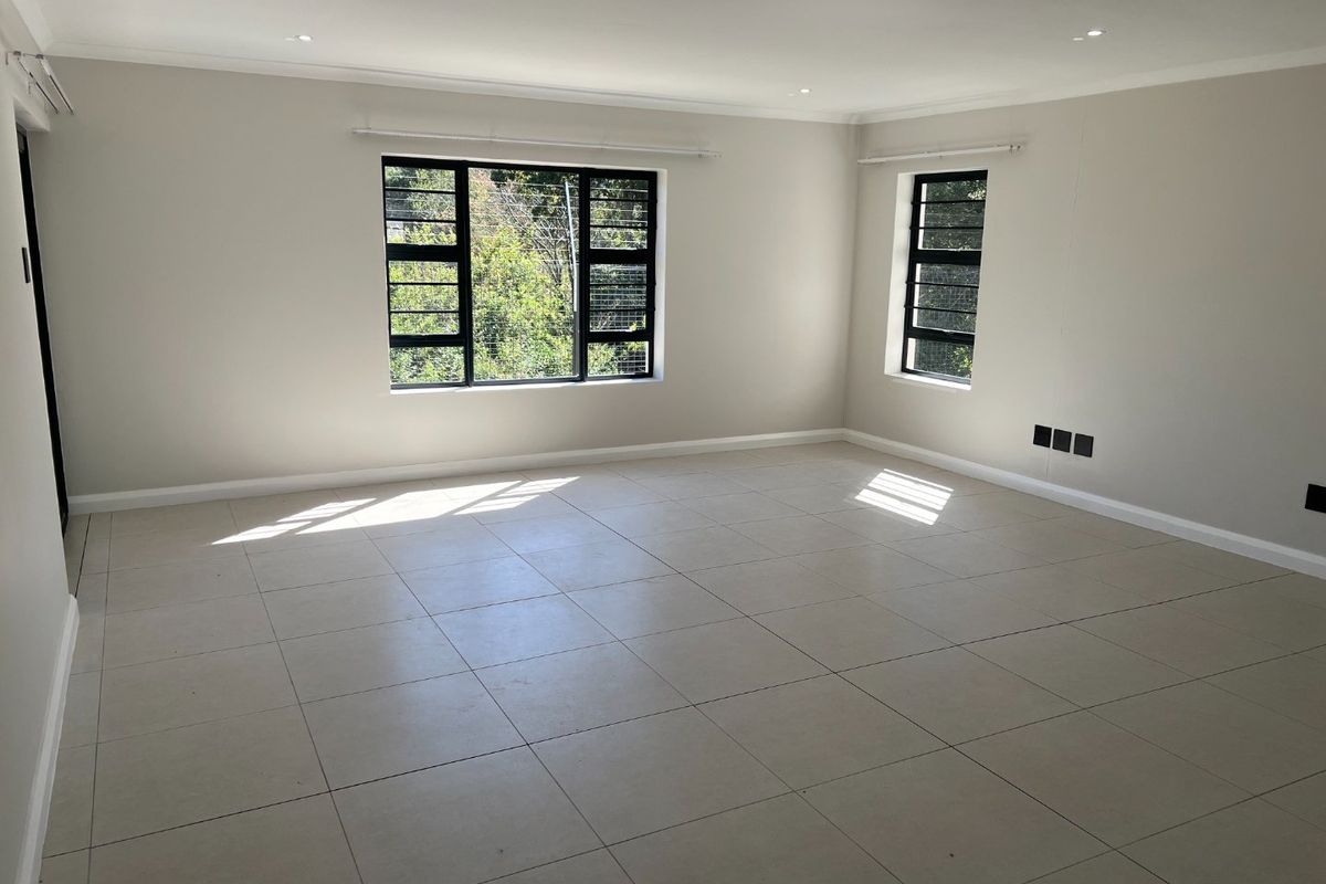 A bright, spacious, and empty living area featuring neutral tiled flooring, recessed downlights, and large windows with black frames and security bars.