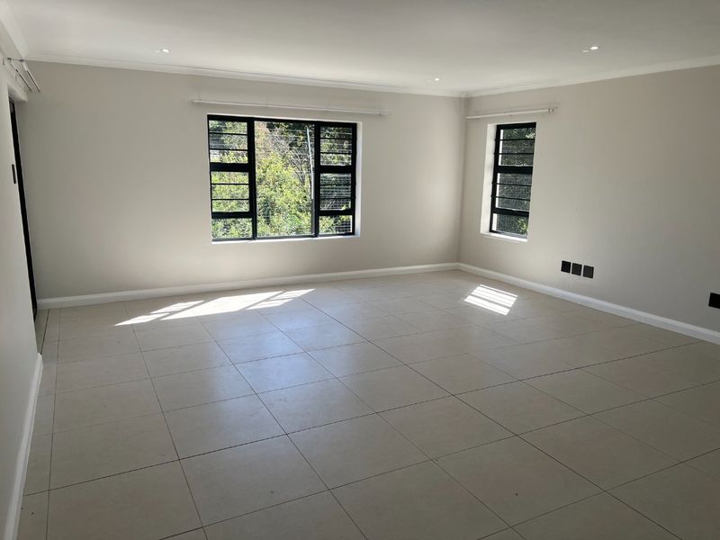 A bright, spacious, and empty living area featuring neutral tiled flooring, recessed downlights, and large windows with black frames and security bars.
