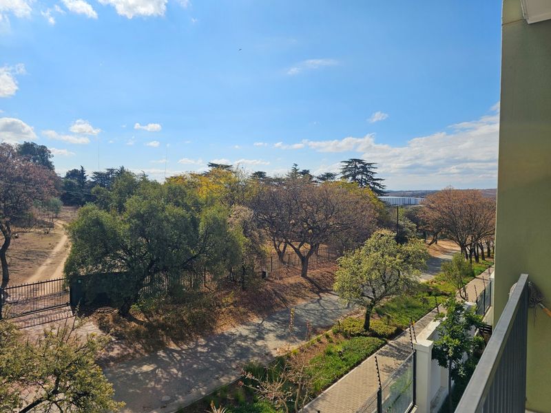 Elevated outdoor view overlooking a landscaped garden area, paved pathways, and mature trees from a balcony perspective.