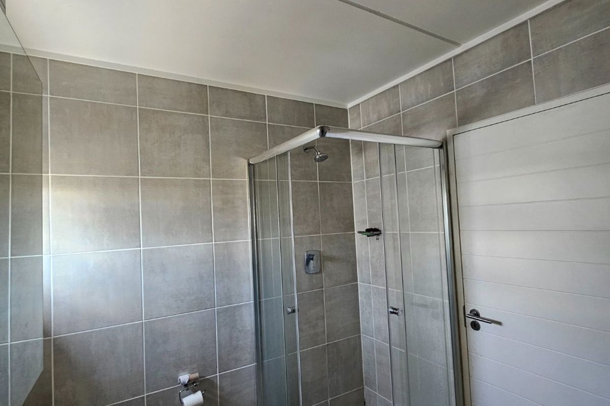 A functional bathroom featuring a corner shower enclosure, pedestal washbasin, toilet, and neutral grey wall and floor tiling.