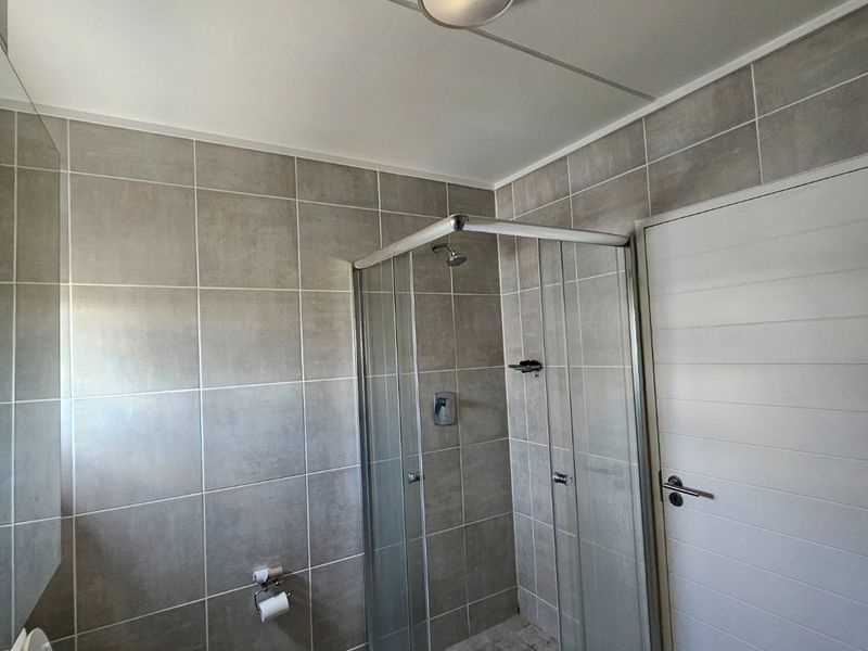 A functional bathroom featuring a corner shower enclosure, pedestal washbasin, toilet, and neutral grey wall and floor tiling.