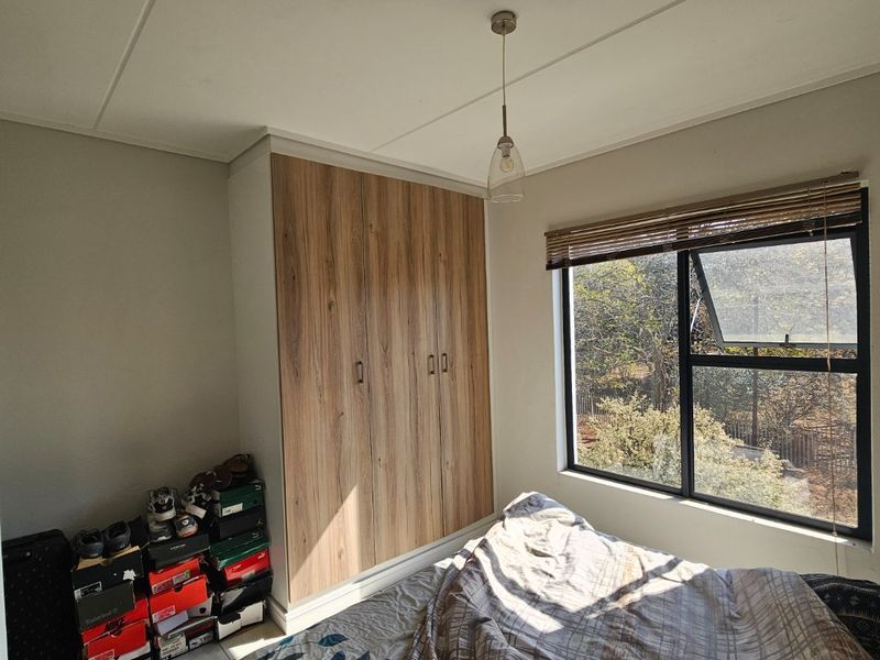 A well-lit bedroom featuring built-in wood-finish wardrobes, tiled flooring, and a large window with venetian blinds overlooking the exterior.