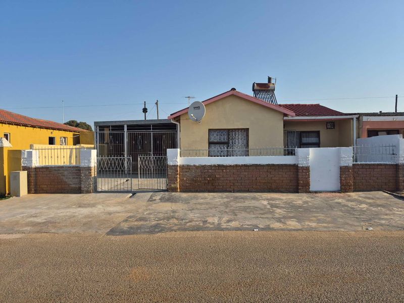 Front elevation of a single-story house featuring a brick boundary wall, metal security gate, and a solar water heater installation.
