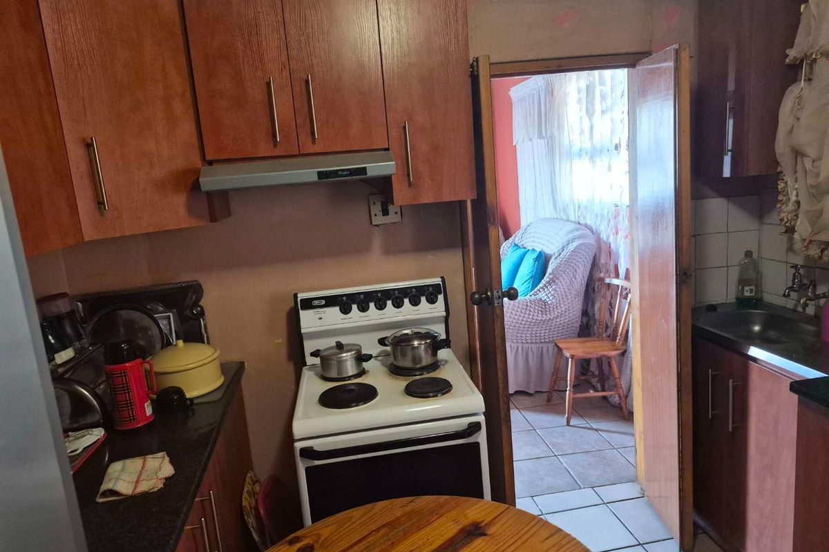 A compact kitchen featuring wood-finish cabinetry, a freestanding stove with an extractor fan, and a stainless steel sink, leading into an adjacent living area.