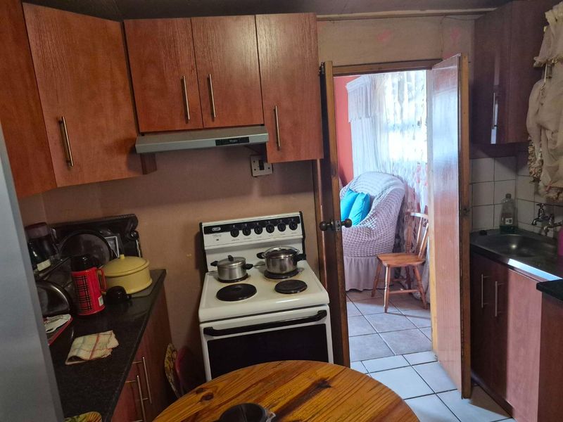A compact kitchen featuring wood-finish cabinetry, a freestanding stove with an extractor fan, and a stainless steel sink, leading into an adjacent living area.