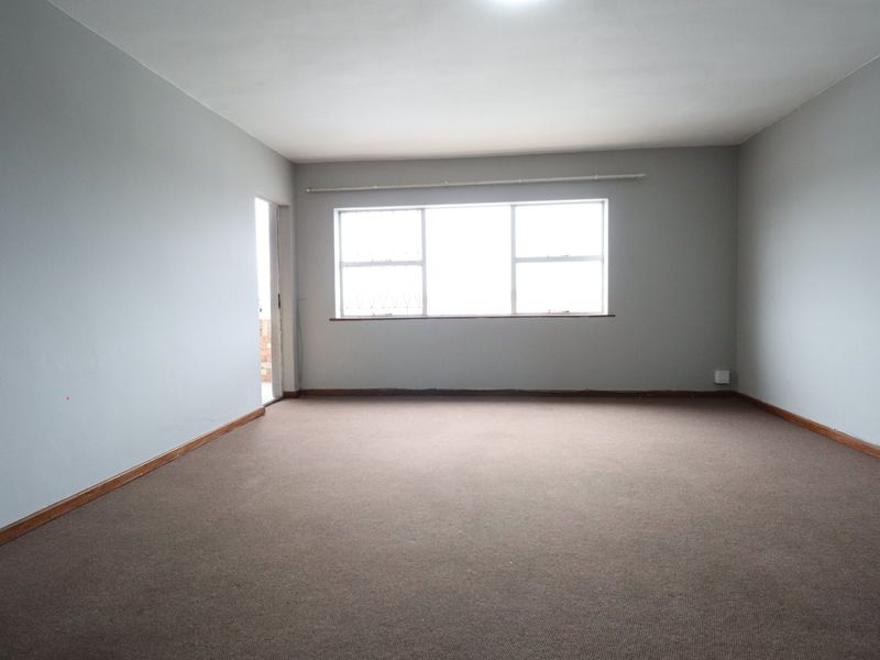 A spacious, empty living room featuring neutral wall paint, carpet flooring, and a large window with security bars.