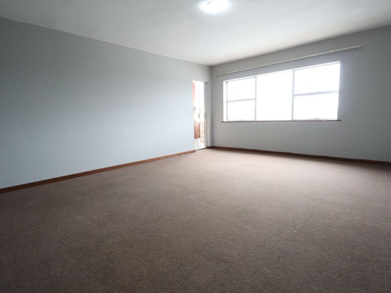 A spacious, empty living room featuring neutral walls, carpeted flooring, and a large window providing natural light.