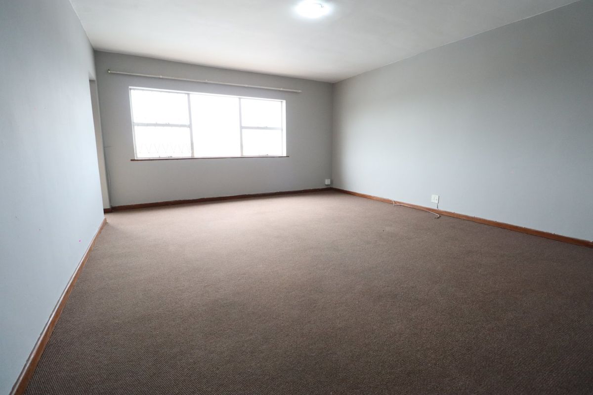 A spacious, empty living room featuring neutral grey walls, carpeted flooring, and a large window providing natural light.