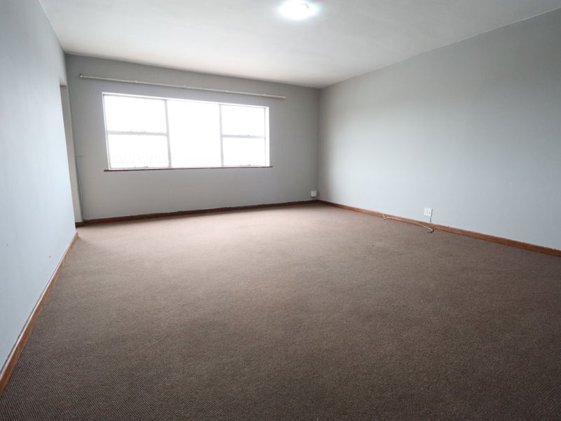 A spacious, empty living room featuring neutral grey walls, carpeted flooring, and a large window providing natural light.
