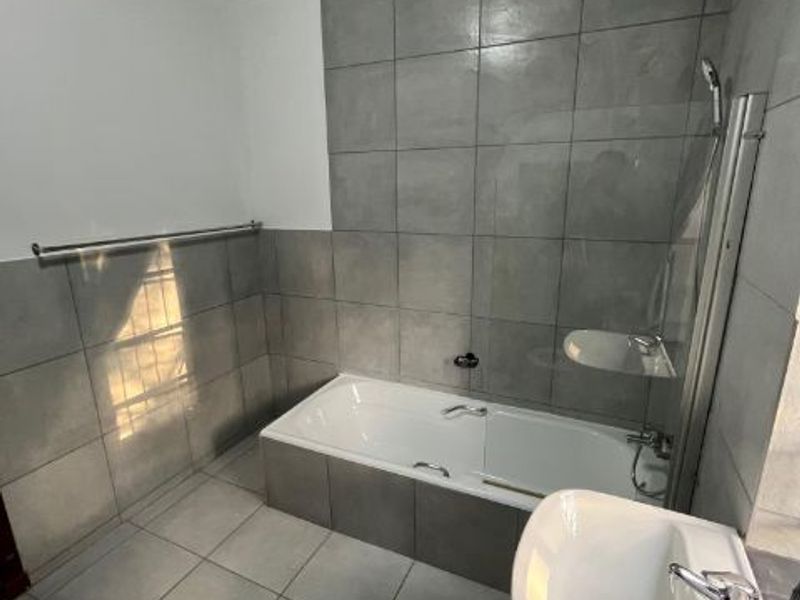 A modern bathroom featuring grey floor-to-ceiling wall tiles, a built-in bathtub with a glass shower screen, and a pedestal basin.