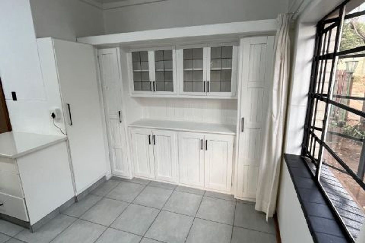 A bright, clean kitchen area featuring white built-in cabinetry with glass-fronted display units, tiled flooring, and large steel-framed windows.