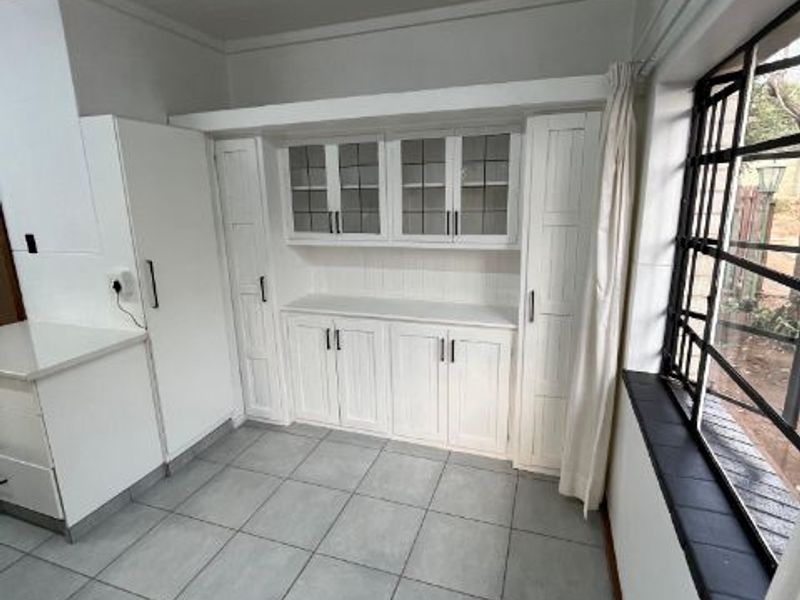 A bright, clean kitchen area featuring white built-in cabinetry with glass-fronted display units, tiled flooring, and large steel-framed windows.